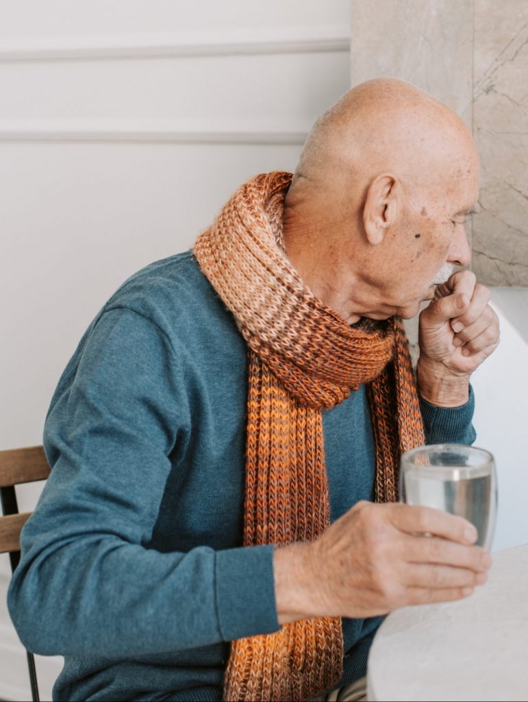 A man coughs into his hand. He is wearing an orange scarf and holding a glass of water in his other hand.